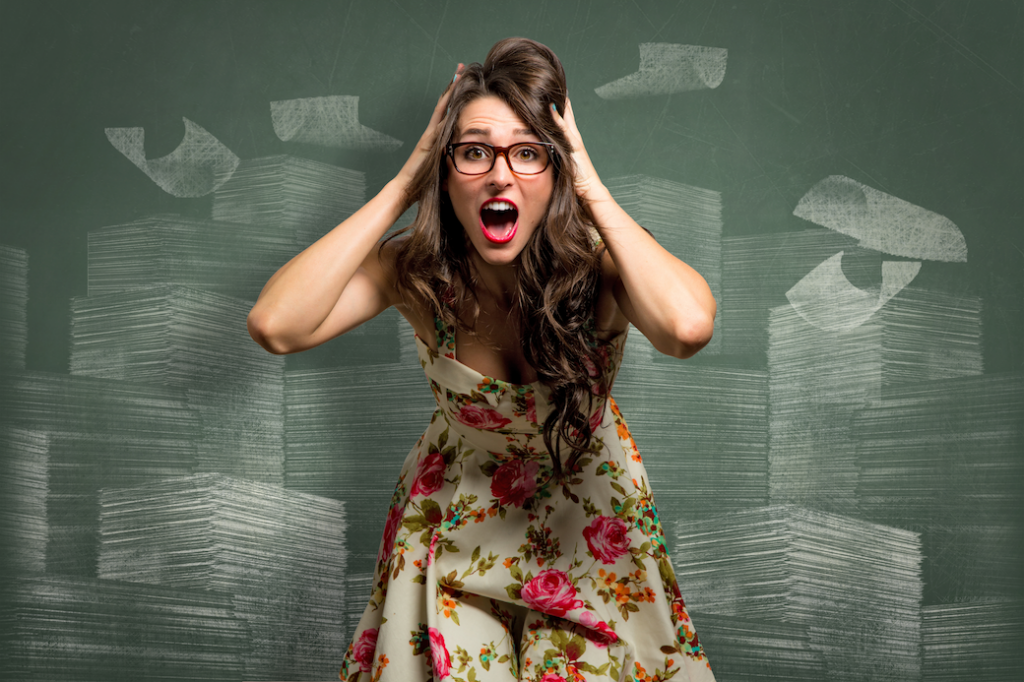 Teacher standing in front of a chalkboard wearing a dress and glasses holding her head with a shocked expression on her face.