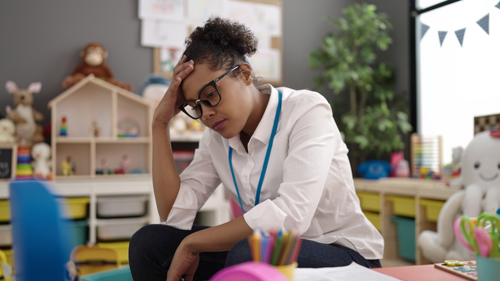 A teacher sitting in and empty classroom holding her head in her hand